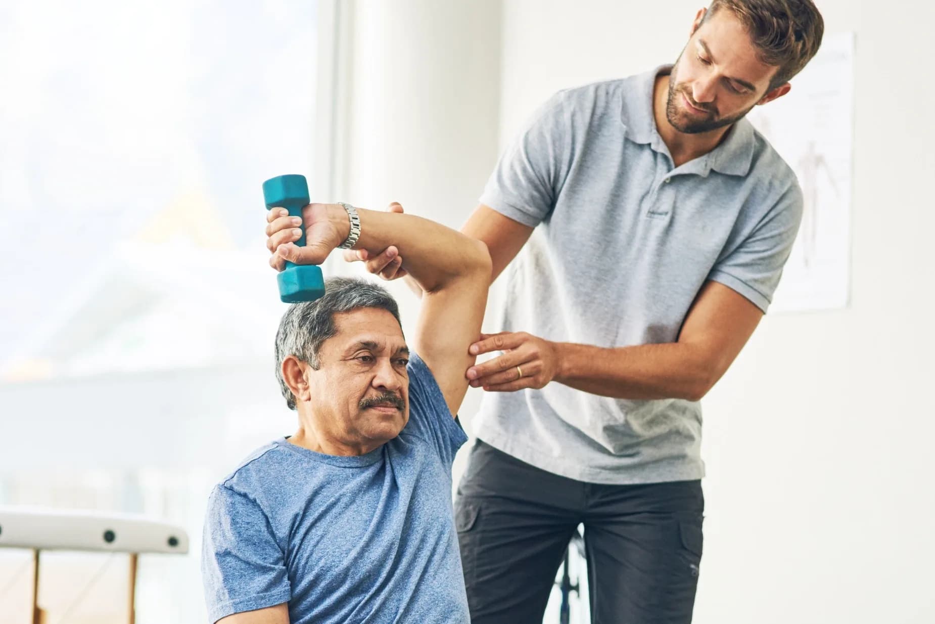 A therapist supports an older man's elbow as he lifts a light dumbbell overhead