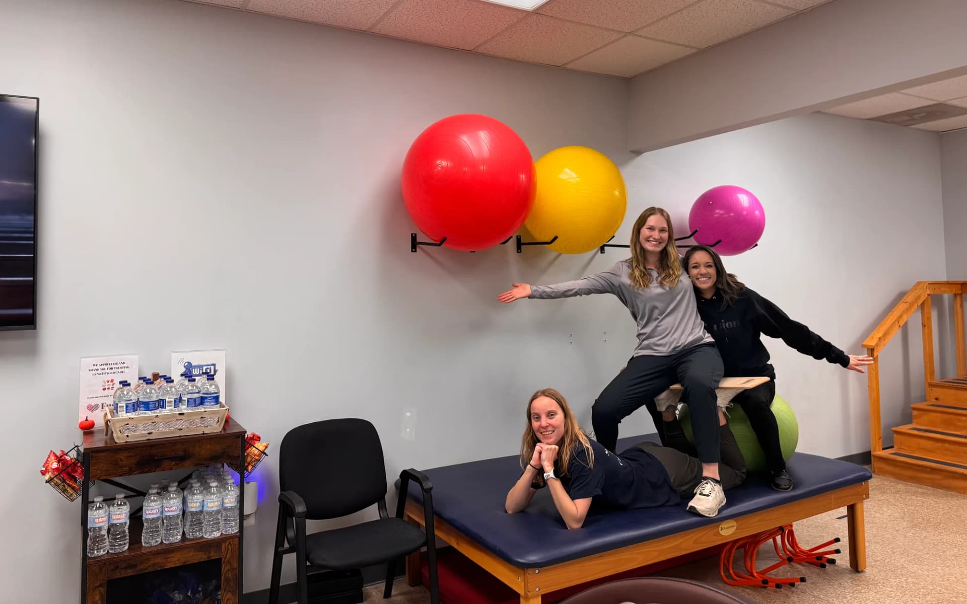 Three people pose playfully on a treatment table and a balance board beneath a wall of exercise balls in a Fusion clinic.